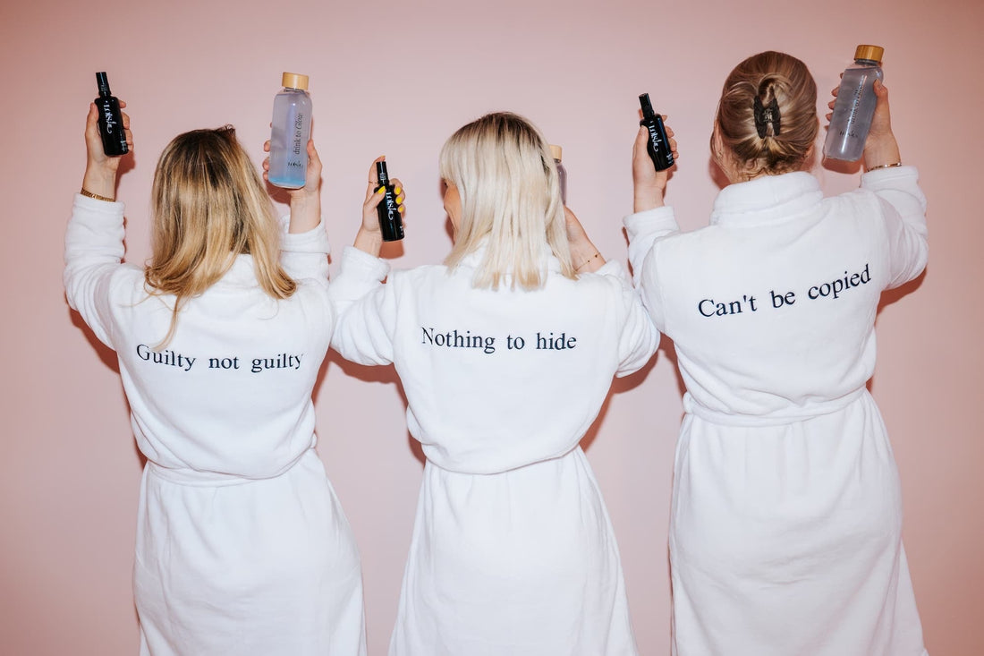 Tre kvinder i hvide badekåber set bagfra, broderet med 'Guilty not guilty', 'Nothing to hide' og 'Can't be copied', holder Loïs Lee produkter op