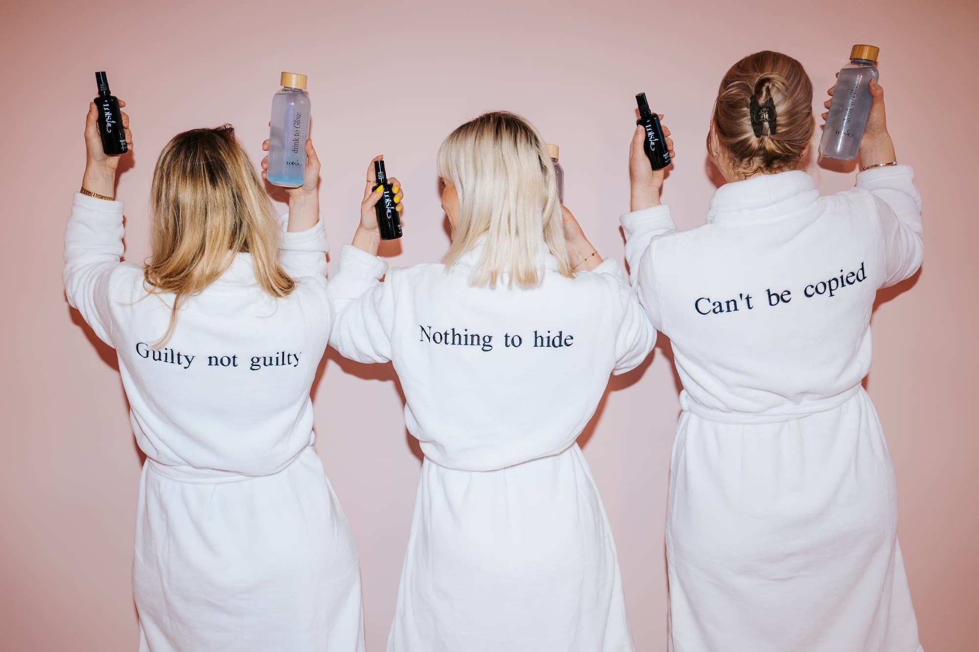 Tre kvinder i hvide badekåber set bagfra, broderet med 'Guilty not guilty', 'Nothing to hide' og 'Can't be copied', holder Loïs Lee produkter op