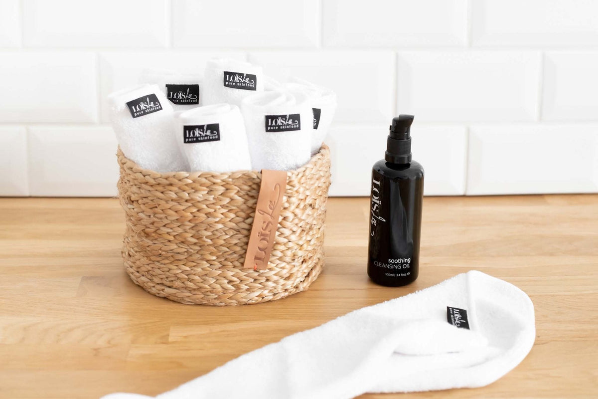 Soothing Cleansing Oil ved siden af en kurv med hvide vaskeklude på en bordplade af træ